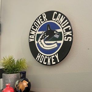 Puede incluir: Un letrero redondo de madera negra con las palabras "VANCOUVER CANUCKS HOCKEY" en blanco. El centro presenta el logotipo del equipo en azul, verde y blanco. El letrero está montado en una pared gris.
