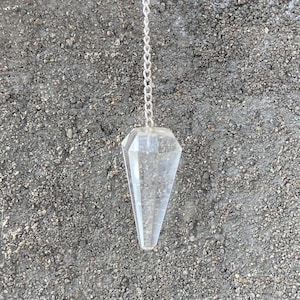 May include: A clear crystal pendulum with a silver chain hangs against a textured grey background. The pendulum is faceted and tapers to a point. The chain is attached to the top of the crystal.