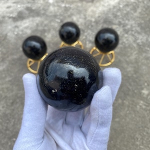 Op de afbeelding: Een grote, gepolijste zwarte bol wordt vastgehouden in een gehandschoende hand, met drie kleinere bollen op goudkleurige standaards op de achtergrond. De bollen hebben een glanzend, reflecterend oppervlak en de achtergrond is neutraal grijs.