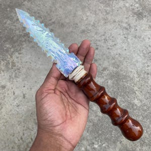 Puede incluir: Una daga decorativa con una hoja clara e iridiscente y un mango de madera marrón oscuro. La hoja tiene un borde dentado y está envuelta con hilo blanco cerca del mango. El mango tiene un diseño similar al bambú.
