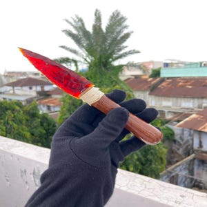 Puede incluir: Una punta de lanza hecha a mano con una hoja roja translúcida y un mango de madera. La hoja está unida al mango con un cordón blanco. La punta de lanza se sostiene en una mano enguantada de negro.