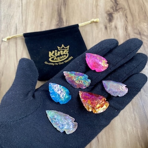 May include: A hand wearing a black glove holds several iridescent, arrowhead-shaped crystals in shades of pink, blue, and gold. A black velvet pouch with gold text that reads "King Agate, Quality is Our Identity" is in the background.
