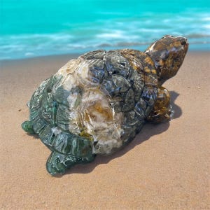May include: A detailed sculpture of a turtle with a textured shell, in shades of green, brown, and white. The turtle is resting on a sandy beach with the ocean in the background. The sculpture is a decorative item.