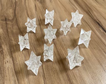 Puede incluir: Una colección de estrellas Merkaba de cristal transparente y facetado. Estas formas geométricas están dispuestas sobre una superficie de madera, mostrando su intrincado diseño de múltiples puntas. Los cristales son transparentes.