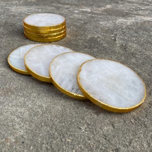 May include: Set of white stone coasters with gold-painted edges. The coasters are round and have a polished finish. A stack of coasters is also visible, showcasing the gold trim.