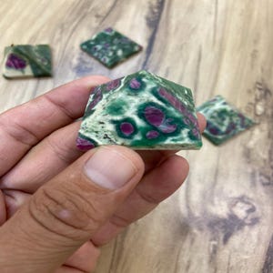 May include: A hand holding a green and pink pyramid-shaped stone. The stone has a marbled appearance with a mix of green and pink hues. Several other similar stones are visible in the background.
