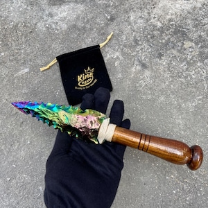 May include: A decorative spearhead with a multi-coloured, iridescent blade and a wooden handle. The handle is wrapped with twine near the blade. A black velvet bag with the text "King Agate" is in the background.
