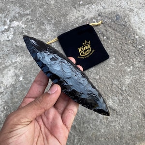 May include: A polished, black obsidian arrowhead with a sharp point, held in a hand. A black velvet pouch with gold drawstring and the text "King Agate" is in the background. The arrowhead is approximately 7 inches long.