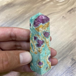May include: A rectangular, polished gemstone with a teal base colour and vibrant magenta inclusions. The stone has a faceted top and a unique pattern of colours and textures. The gemstone is held in a hand.