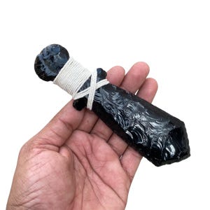Puede incluir: Una daga de obsidiana negra con una superficie texturizada. El mango está envuelto con hilo blanco. La daga tiene un pomo redondeado y una hoja puntiaguda.