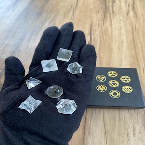 Könnte beinhalten: Ein schwarzer Handschuh hält verschiedene klare Kristallformen, darunter eine Pyramide, ein Quadrat, ein Stern und eine Kugel. Eine kleine schwarze Schachtel mit goldenen geometrischen Designs ist ebenfalls vorhanden. Die Kristalle werden auf einer Holzoberfläche präsentiert.