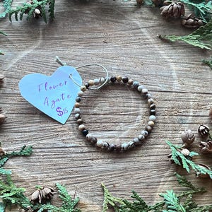 Puede incluir: Un brazalete de ágata de flor marrón, blanco y negro sobre una superficie de madera con follaje verde. El brazalete está hecho de pequeñas cuentas redondas. Una etiqueta en forma de corazón con el texto "Flower Agate $15" está en la imagen.