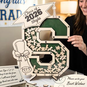 May include: A wooden graduation guest book in the shape of the letter "S", with a green interior and a banner that reads "Class of 2026 Shyla". Includes a box for guests to sign and a sign that says "Please write your Best Wishes".