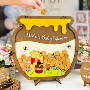 Può includere: Libro degli ospiti per baby shower in legno a forma di vaso di miele, con la scritta "Alida's Baby Shower". Il design include un'illustrazione di Winnie the Pooh, api e forme a nido d'ape. Una mano sta mettendo un gettone di legno.