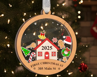May include: A round, wooden Christmas ornament with a clear center, featuring a house design with the year 2025. The ornament includes festive graphics and the text "FIRST CHRISTMAS AT 305 Main St". It hangs from a silver ribbon.