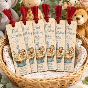 May include: Six rectangular wooden bookmarks with red fabric tassels, each featuring a blue cupcake illustration with a monkey, the text First Birthday Helen, a large number 1, and the date 09.09.2026, all arranged inside a woven wicker basket.