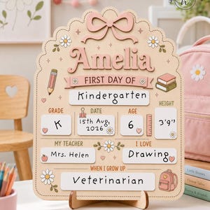 May include: A light-colored wooden sign for the first day of school featuring the name Amelia, a pink bow, and daisy illustrations. It includes fields for grade, date, age, height, teacher, interests, and future career, with text reading 15th Aug, 2026.