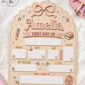 May include: A light-colored wooden sign with a scalloped edge, featuring a pink bow at the top and the name Amelia. It includes blank spaces for school details like grade, date, age, height, teacher, interests, and future goals, decorated with daisies, a pencil, books, and a backpack.