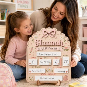 May include: A light-colored wooden sign with a scalloped edge and a pink bow at the top. The text reads "Hannah" and "LAST DAY OF". It includes fields for "Kindergarten", "K", "May 23, 2026", "6", "3'9"", "Mrs. Carter", "Drawing", and "Veterinarian".