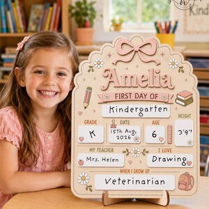 May include: A wooden first day of school sign featuring a scalloped edge, a pink bow, and floral accents. The sign displays the name Amelia, grade K, date 15th Aug, 2026, age 6, height 3'9", teacher Mrs. Helen, interest in drawing, and career as a veterinarian.