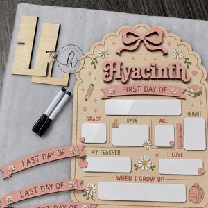 May include: A wooden school milestone board featuring the name Hyacinth, a pink bow, and daisy accents. It includes blank white spaces for grade, date, age, height, teacher, interests, and future goals, plus two markers and interchangeable banners.