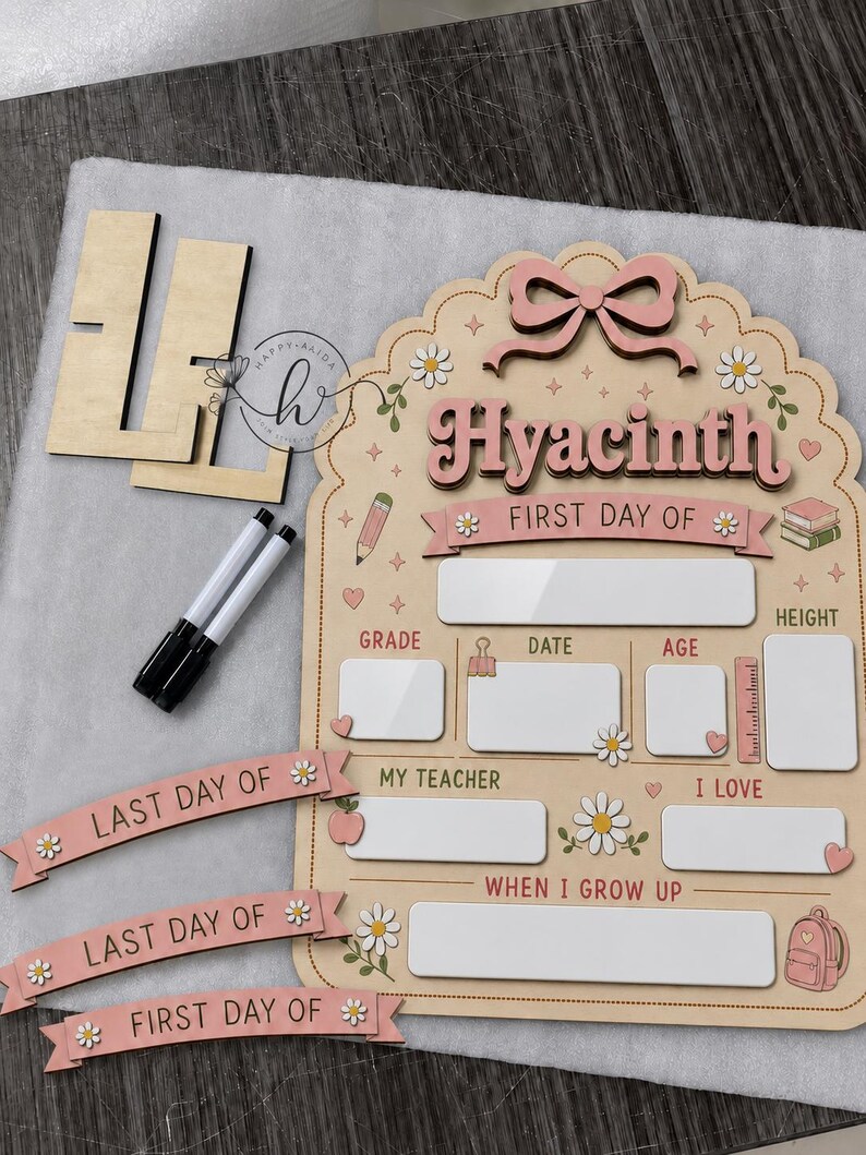 May include: A wooden school milestone board featuring the name Hyacinth, a pink bow, and daisy accents. It includes blank white spaces for grade, date, age, height, teacher, interests, and future goals, plus two markers and interchangeable banners.