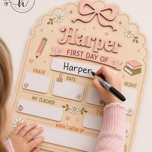 May include: A light-colored wooden sign with a scalloped edge, featuring a pink bow, daisy illustrations, and text including "Harper," "FIRST DAY OF," "GRADE," "DATE," "HEIGHT," "MY TEACHER," and "WHEN I GROW UP," with blank spaces for writing.