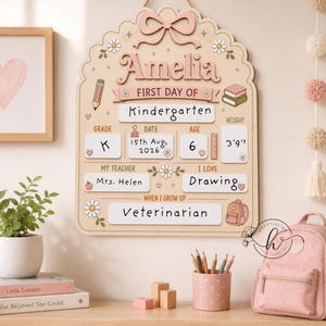 May include: A light-colored wooden wall sign with a scalloped top and a pink bow, featuring the name Amelia and the text First Day of Kindergarten. It includes sections for grade, date, age, height of 3'9", teacher, interests, and future career.