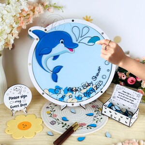May include: A baby shower guest book alternative featuring a blue dolphin design. The round sign has the text "Alida's Baby Shower 09.09.2025". Includes a small sign that says "Please sign my Guest Book". A box of blue and white tokens is also included.