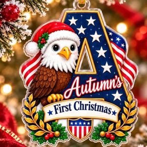 May include: A clear acrylic Christmas ornament shaped like the letter A, featuring a bald eagle wearing a Santa hat, American flag motifs, and the text Autumn's First Christmas. The design includes gold laurel leaves and a small shield emblem.