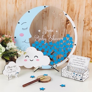 Könnte beinhalten: Ein blaues und weißes Mond- und Wolken-Babyparty-Gästebuch mit einem Schild, auf dem "Bitte unterschreiben Sie mein Gästebuch" steht. Das Gästebuch besteht aus klarem Acryl und hat einen Schlitz, in den die Gäste ihre Glückwünsche einwerfen können. Das Schild ist aus Holz und hat einen weißen Hintergrund mit schwarzem Text. Der Mond und die Wolke sind mit Sternen und dem Text "Jame Baby Shower" verziert.