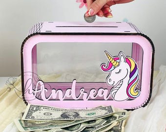 Può includere: Un salvadanaio in legno rosa con un pannello frontale trasparente e una grafica di unicorno. La scatola ha il nome "Andrea" in corsivo. Una mano sta inserendo una moneta nella fessura. Sono visibili diverse banconote.