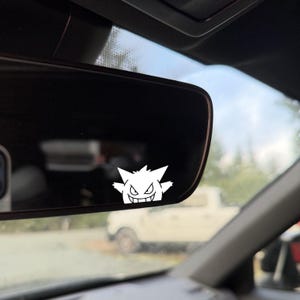 May include: A white vinyl sticker of a cartoon ghost character with a spiky head and a sinister grin is affixed to a car's rearview mirror. The sticker is in sharp focus, contrasting with the blurred background of a vehicle and trees.