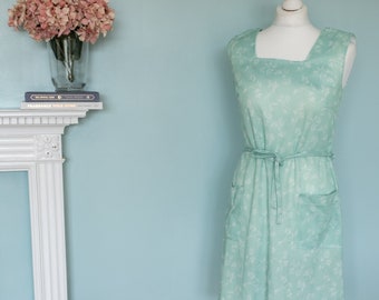 May include: A light green dress with a white floral pattern, a tie waist, and two pockets. The dress is on a white dress form with a white stand. A brown tufted ottoman is in the foreground. The background is a light blue wall.
