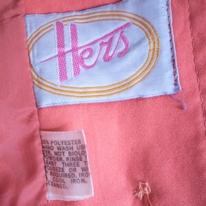 May include: A white tag with the word "Hers" embroidered in pink and a gold oval around it. The tag is attached to a coral pink fabric garment. The tag also includes care instructions for the garment, including hand washing and cool ironing.