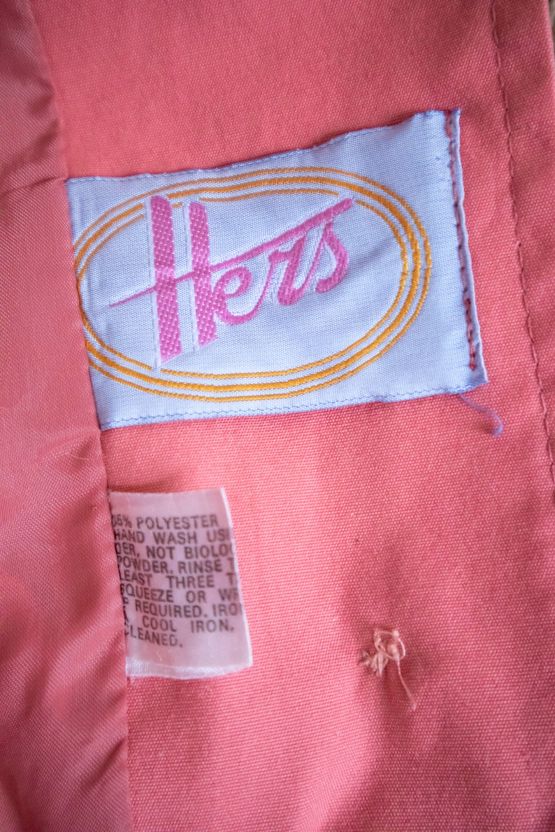 May include: A white tag with the word "Hers" embroidered in pink and a gold oval around it. The tag is attached to a coral pink fabric garment. The tag also includes care instructions for the garment, including hand washing and cool ironing.
