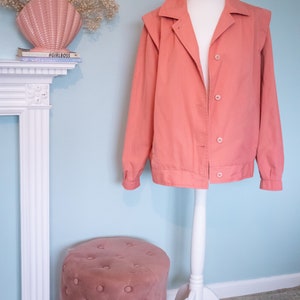 May include: A salmon-colored jacket with a white stand collar and button closure. The jacket is on a white mannequin with a white stand. A pink velvet footstool is in the foreground.