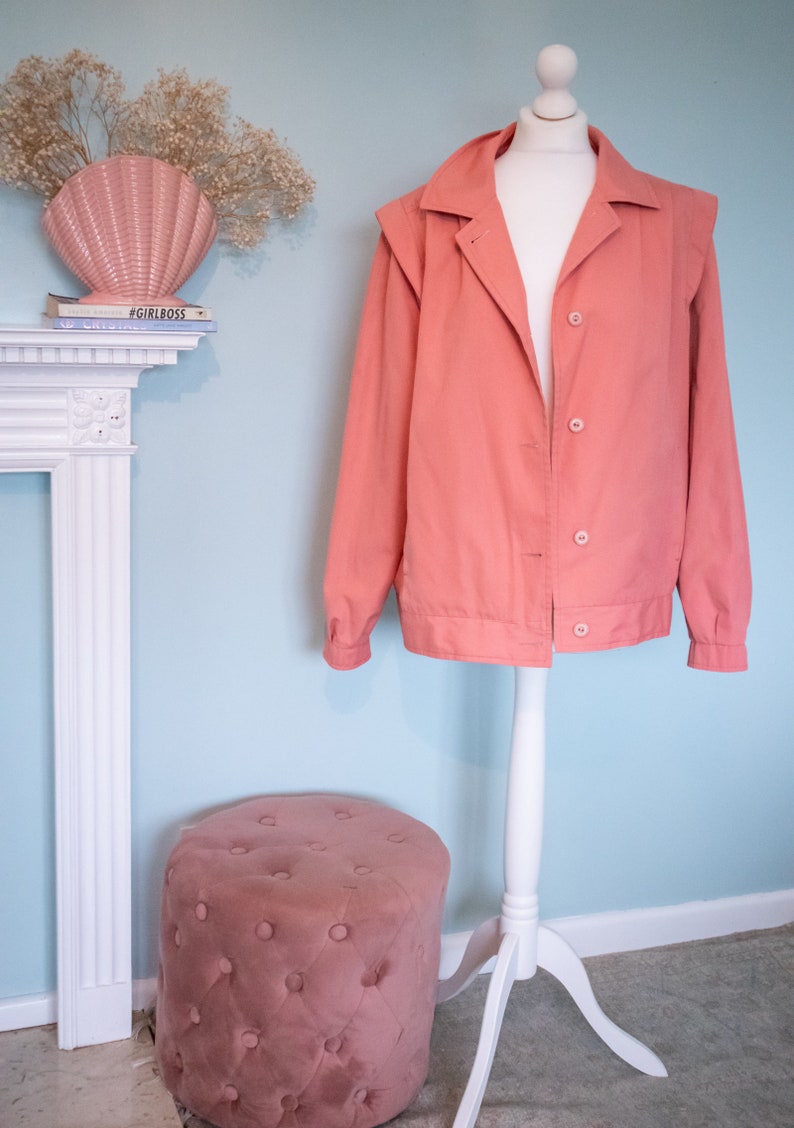 May include: A salmon-colored jacket with a white stand collar and button closure. The jacket is on a white mannequin with a white stand. A pink velvet footstool is in the foreground.