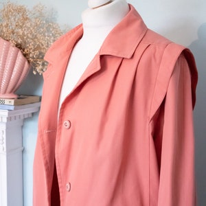 May include: A salmon pink long-sleeved jacket with a button-up front and a collar. The jacket has a slightly oversized fit and features a pleated detail on the back.