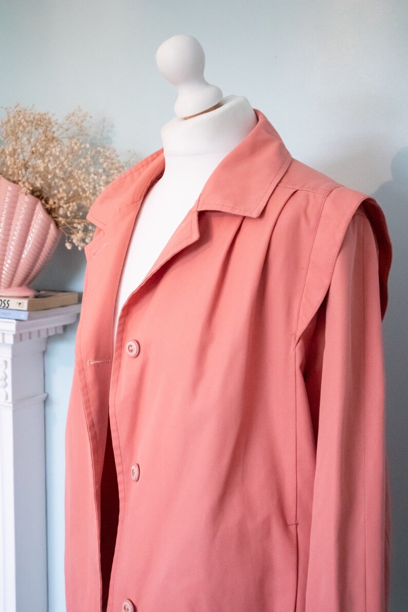 May include: A salmon pink long-sleeved jacket with a button-up front and a collar. The jacket has a slightly oversized fit and features a pleated detail on the back.