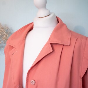 May include: A coral pink jacket with a button closure and a collar. The jacket has a pleated design on the front and a slightly puffed shoulder.