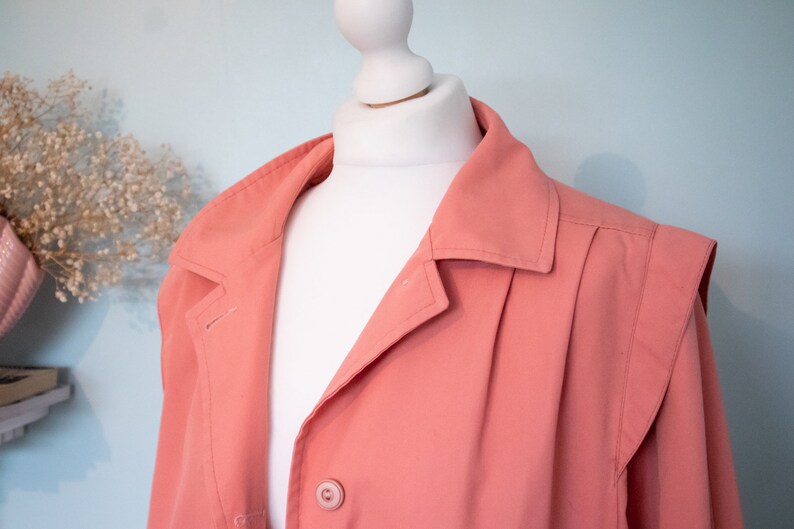 May include: A coral pink jacket with a button closure and a collar. The jacket has a pleated design on the front and a slightly puffed shoulder.