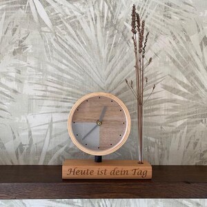 Könnte beinhalten: Eine Holz-Schreibtischuhr mit hellem Holzrahmen und grauem und holzfarbenem Zifferblatt. Die Uhr steht auf einem Holzsockel mit dem Text "Heute ist dein Tag". Neben der Uhr steht eine kleine Glasvase mit getrockneten Pflanzen.