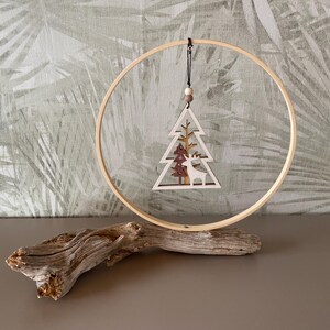 Könnte beinhalten: Ein hölzerner Weihnachtsschmuck mit einem lasergeschnittenen Design eines Weihnachtsbaums und eines Hirsches, der in einem Holzreifen aufgehängt ist. Der Schmuck wird auf einem Stück Treibholz präsentiert und schafft eine rustikale Weihnachtsdekoration.