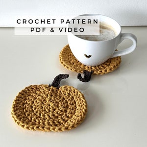 Może przedstawiać: Dwa dzianinowe podkładki pod kubki w kształcie dyni w kolorze musztardowym. Podkładki znajdują się na białym tle. Tekst "CROCHET PATTERN PDF & VIDEO" jest wyświetlany nad podkładkami.