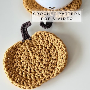 Peut inclure: Un dessous de verre en forme de citrouille crocheté de couleur jaune moutarde avec une tige brune. Le dessous de verre est posé sur une surface blanche. Le texte "CROCHET PATTERN PDF & VIDEO" est affiché au-dessus du dessous de verre.