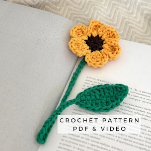 May include: A crochet sunflower bookmark with a green stem and leaves. The sunflower is yellow with a brown center. The bookmark is laying on a book with the text "CROCHET PATTERN PDF & VIDEO" on the page.