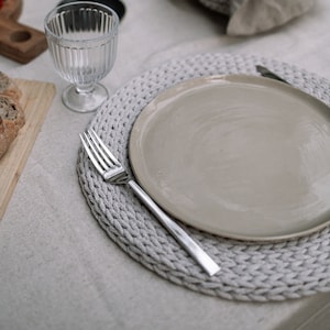 May include: A close-up of a table setting with a white tablecloth, a gray crocheted placemat, a beige plate, a silver fork, and a clear glass.
