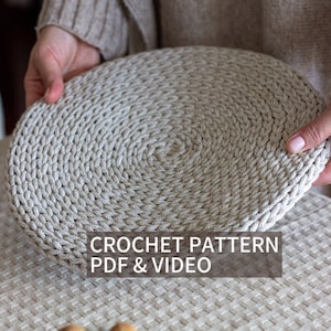 May include: A close-up of a crocheted round coaster in a light beige color. The coaster is being held by a hand. The text "CROCHET PATTERN PDF & VIDEO" is displayed below the coaster.