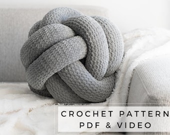 Puede incluir: Un cojín de nudo tejido en color gris sobre un sofá blanco y gris. El texto "CROCHET PATTERN PDF & VIDEO" se muestra debajo del cojín.